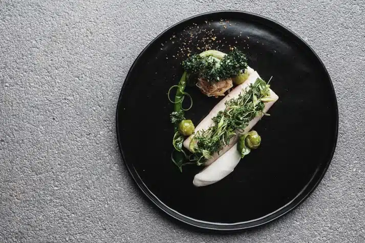 Ein Fischfilet mit grünem Gemüse und Kräutern, angerichtet auf einem schwarzen Teller.