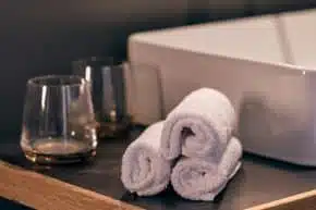 Trois serviettes blanches roulées sont posées sur le comptoir de l'évier à côté de deux verres propres dans la salle de bains de l'Hôtel LOISIUM Champagne.