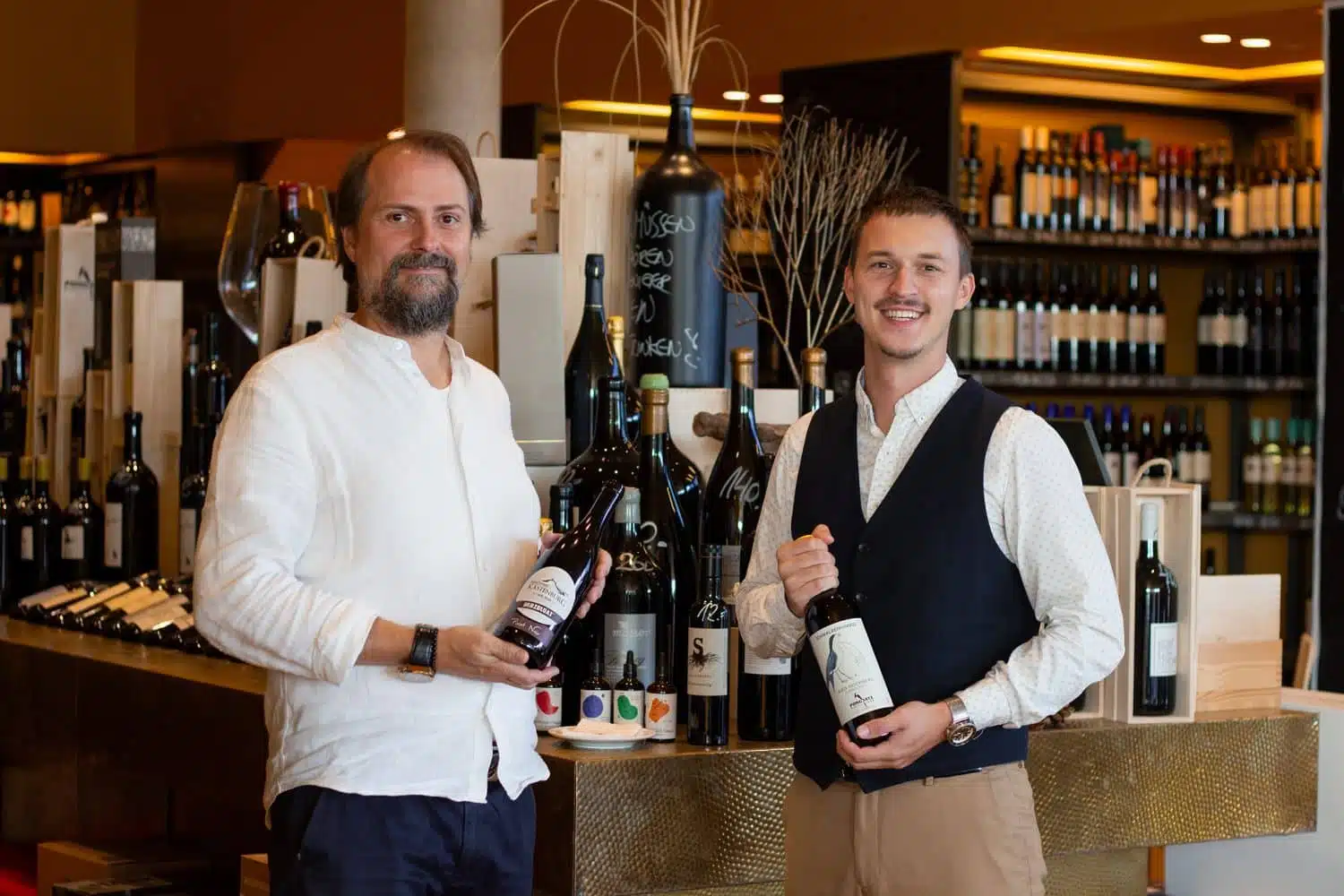 Zwei Mitarbeiter posieren mit Weinflaschen in der Vinothek