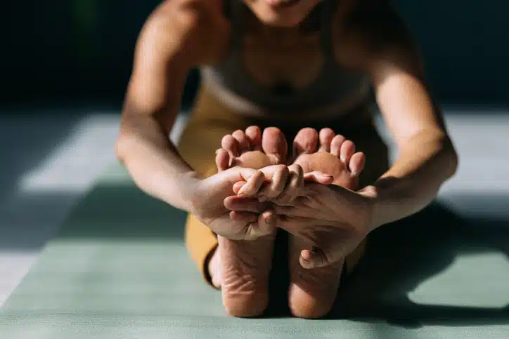 Nahaufnahme einer Person in einer sitzenden Vorwärtsbeuge-Yoga-Position, die mit den Händen die Füße umklammert, auf einer Yogamatte.