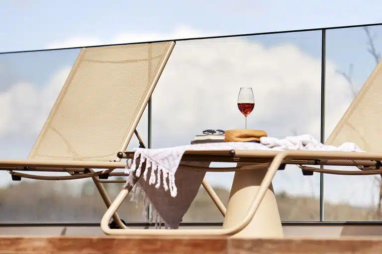 Beige deck chairs on a terrace with a glass of rosé wine, books, straw hat, and towel in front of glass railing and blue sky.