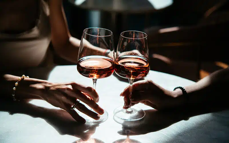Zwei Personen stoßen mit Gläsern Roséwein an, im warmen Licht auf einem runden Tisch.