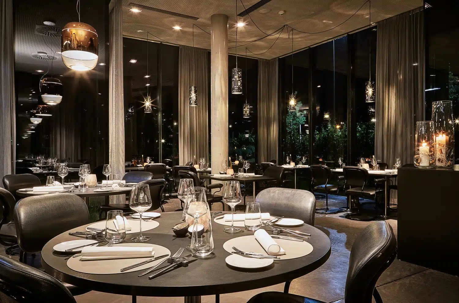 Elegant gedecktes Restaurant im LOISIUM Langenlois mit runden Tischen, stilvollen Lampen und Blick auf den nächtlichen Garten durch bodentiefe Fenster.